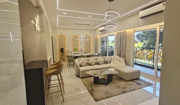 Spacious open-concept living and dining area in the Nambiar District 25 model flat, featuring a luxury L-shaped sofa, marble coffee table, recessed ceiling LED lighting, and arched wall niches.