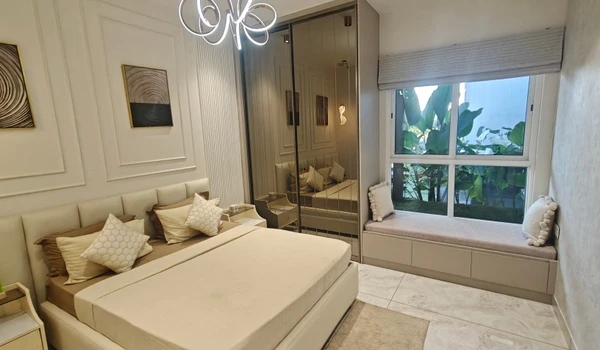 Luxurious master bedroom at Nambiar District 25 model flat featuring a king-sized bed with plush neutral-toned bedding, wall molding accents, a modern gold chandelier, and a cozy window seat overlooking lush greenery.