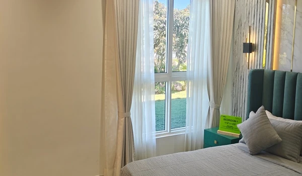 Modern bedroom (Bedroom 1) in Nambiar District 25 4 BHK model flat, featuring a stylish green upholstered headboard, large window with garden view, and contemporary wall sconce lighting