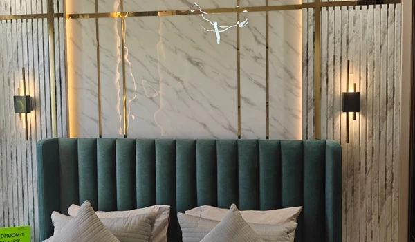 Close-up detail of the luxurious green upholstered headboard in Bedroom 1 of the Nambiar District 25 4 BHK model flat, set against a marble-finish wall with gold accents and modern sconce lighting