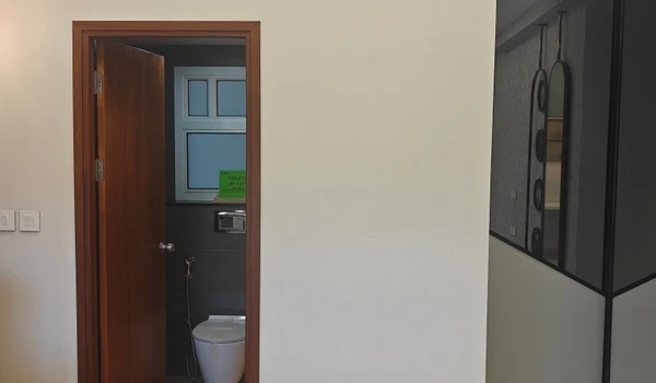 View of the attached bathroom entrance in a Nambiar District 25 4 BHK bedroom, showing a wooden door frame, wall-mounted commode, and modern tiling