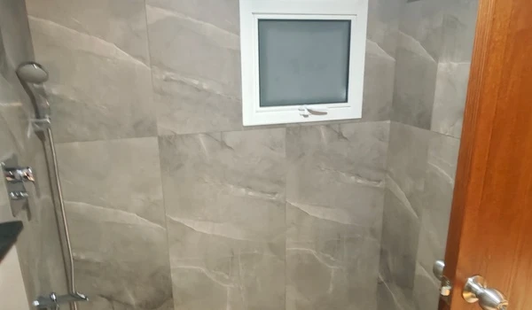 Shower area in the master bathroom of Nambiar District 25 4 BHK model flat, featuring grey stone-finish tiles, premium chrome fixtures, and a ventilation window