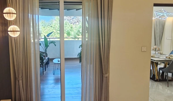 View from the master bedroom through sliding glass doors to the private balcony with outdoor seating, adjacent to the dining area in the Nambiar District 25 4 BHK model flat