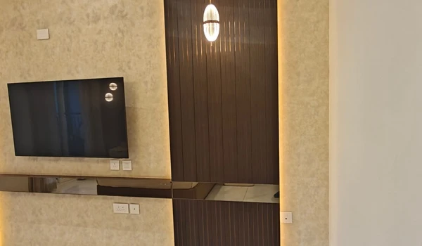 Close-up of the master bedroom TV wall in Nambiar District 25 4 BHK model flat, featuring a textured beige backdrop, ambient LED backlighting, and a dark vertical accent panel with a modern pendant light