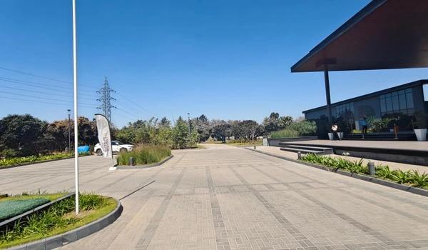 Expansive paved entrance plaza and walkway at Nambiar District 25, bordered by green landscaping and promotional flags under a clear blue sky