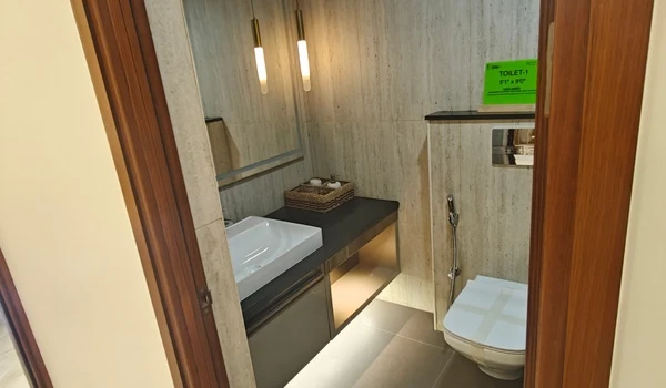 Modern bathroom design at Nambiar District 25 featuring a sleek vanity with a white vessel sink, black countertop, wall-mounted commode, and warm pendant lighting against textured wall tiles
