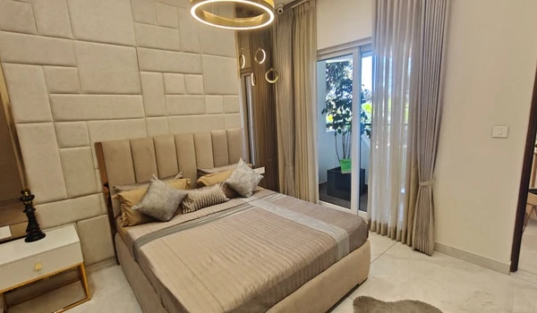 Modern bedroom at Nambiar District 25 model flat featuring a king-sized bed with an upholstered block-pattern headboard, gold circular pendant lighting, and large sliding glass doors opening to a private balcony with greenery