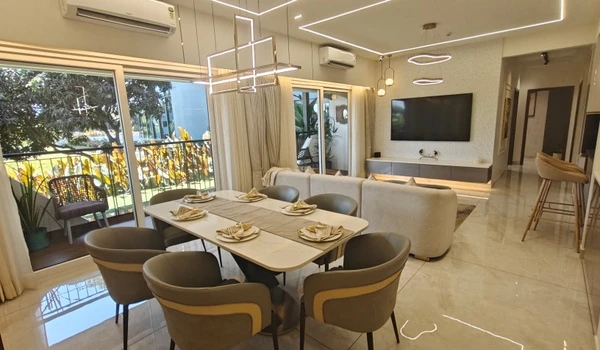 Modern open-plan dining and living area at Nambiar District 25 featuring an 8-seater dining table, luxury L-shaped sofa, wall-mounted TV, and large sliding glass doors leading to a balcony