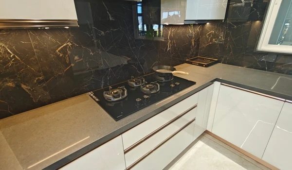 Close-up of a modern modular kitchen at Nambiar District 25, featuring a black marble backsplash with gold veining, a built-in glass hob, and sleek white high-gloss cabinets