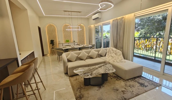 Spacious living room at Nambiar District 25 model flat featuring a luxury L-shaped sofa, marble coffee table, designer pendant lights, and large sliding glass doors opening to a balcony