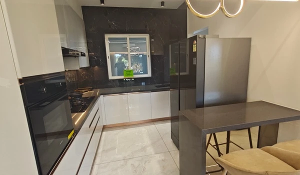 Modern modular kitchen at Nambiar District 25 model flat featuring white high-gloss cabinetry, a black marble backsplash, integrated oven, and a sleek breakfast counter with seating