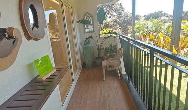 Spacious private balcony at Nambiar District 25 model flat featuring wood-finish flooring, decorative wall art, large planters with tropical greenery, and a cozy wicker chair