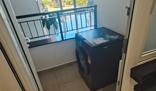 Dedicated utility balcony at Nambiar District 25 model flat, featuring a modern front-load washing machine, anti-skid floor tiles, and a secure metal railing with a view of the project greenery
